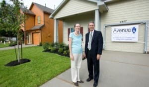 Couple in business attire stand on a driveway in front of a beige house with an Avenue banner celebrating 20 years.