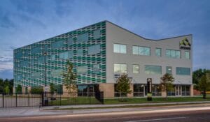 Modern four‑story office building with a teal patterned facade, gray wing, and Avenue sign on the corner beside a street.