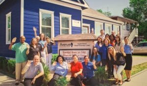 Diverse group of volunteers waving in front of a blue house with an Avenue NeighborhoodWorks HomeOwnership Center sign.