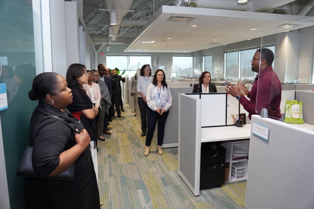 Man in a maroon shirt speaking to a group of colleagues in a modern office reception area.