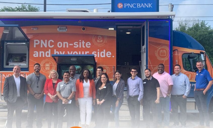Group of diverse employees posing in front of a PNC on-site mobile banking van with orange branding and a PNC logo in the foreground.
