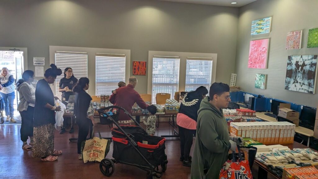 People line up inside a community pantry, shopping at tables piled with boxes and canned goods.