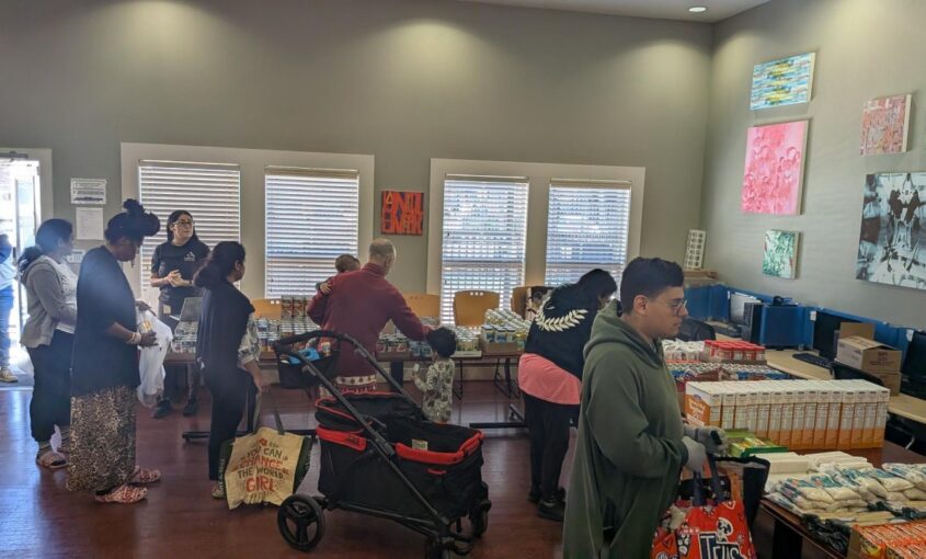 People line up inside a community pantry, shopping at tables piled with boxes and canned goods.