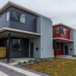 Row of modern two-story townhouses with bold gray, red, blue, and yellow siding and small front yards along a sidewalk and driveway.