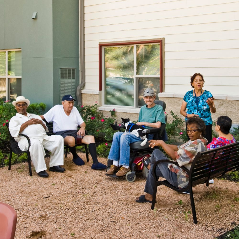 Group of seniors sitting and chatting in a courtyard outside a building, with a woman standing and speaking.