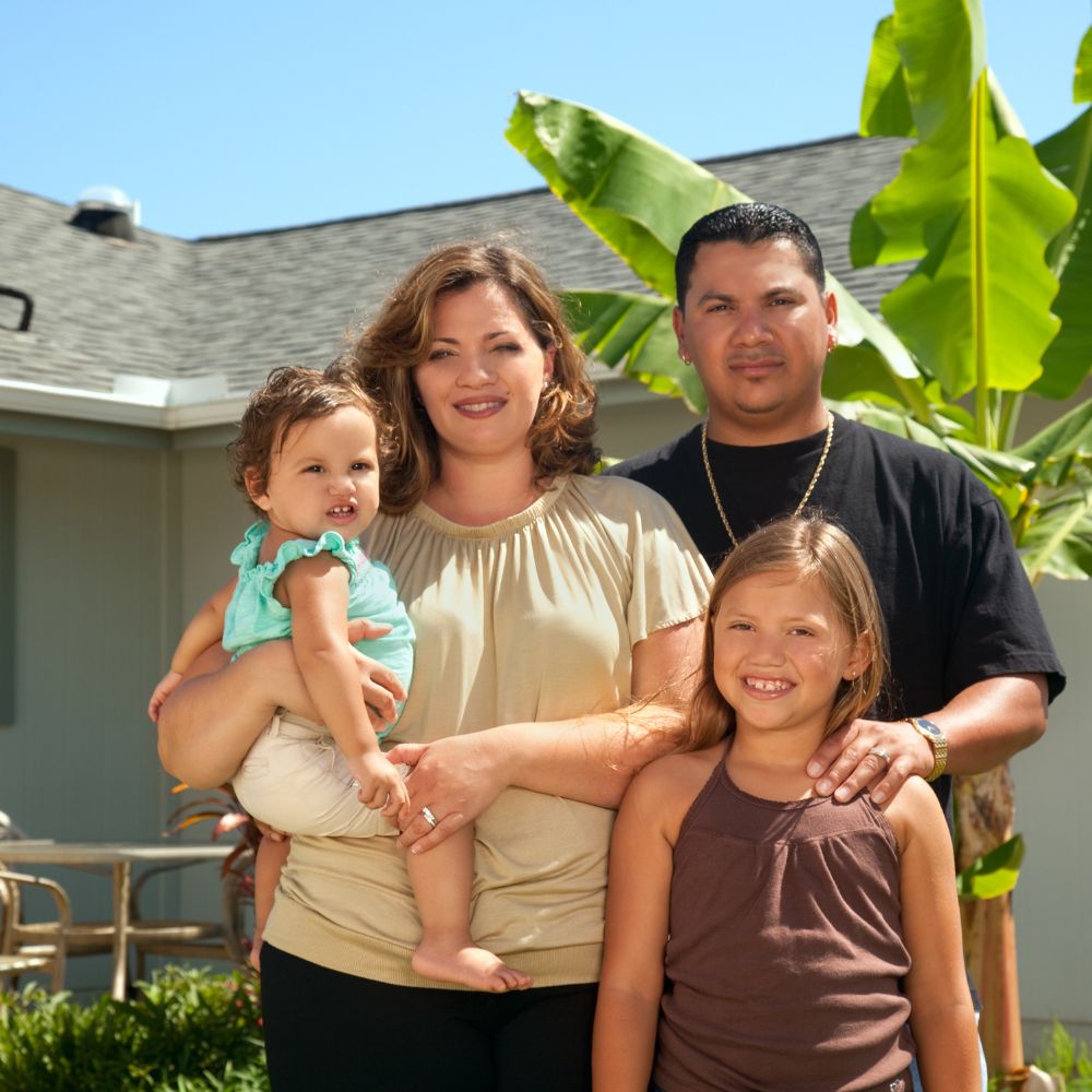 Family of four posing outdoors in a sunny yard; mother holds a toddler, father stands behind with daughter in front.
